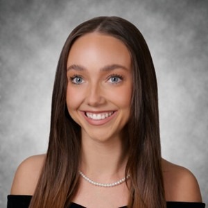Payton Vanden Boom, a young woman with long straight brown hair, smiling in a professional headshot. She is wearing a black off-the-shoulder top and a pearl necklace, posed against a gray studio background.